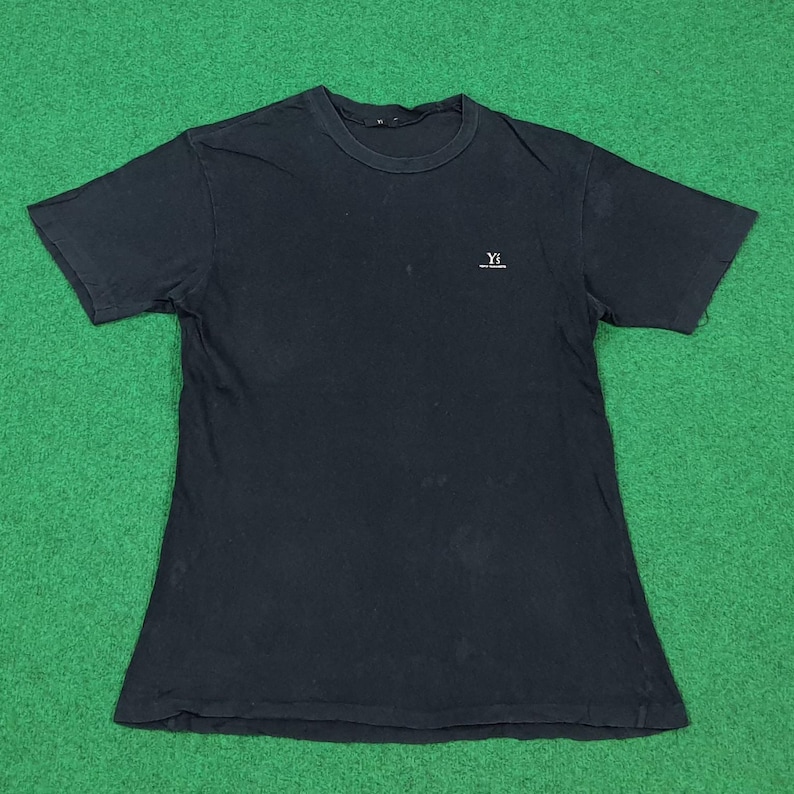 May include: A black short-sleeved t-shirt with a crew neckline. The shirt features a small white logo on the upper left chest area that reads Y's with smaller text underneath. The fabric appears to have a slightly faded, worn-in texture throughout.