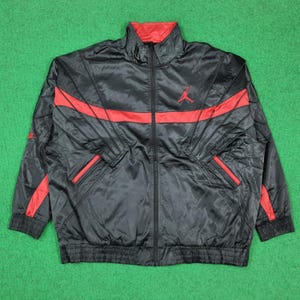 May include: A black track jacket with a red inner collar and red horizontal stripes across the chest and sleeves. The jacket features a full-length front zipper, ribbed cuffs and hem, and a red Jumpman logo on the upper left chest area.