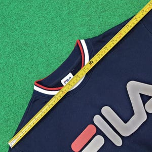 Puede incluir: Sudadera FILA azul marino con cuello a rayas blancas y rojas. El logotipo de FILA se muestra en blanco, rojo y gris. Una cinta m&eacute;trica amarilla se coloca sobre la sudadera, mostrando medidas en cent&iacute;metros.