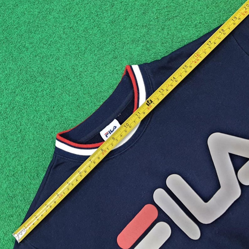 Puede incluir: Sudadera FILA azul marino con cuello a rayas blancas y rojas. El logotipo de FILA se muestra en blanco, rojo y gris. Una cinta m&eacute;trica amarilla se coloca sobre la sudadera, mostrando medidas en cent&iacute;metros.