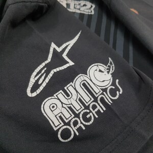 May include: A dark gray t-shirt featuring a white graphic print of a stylized star logo and the text Ryno Organics with a rhinoceros icon. The white ink shows visible cracking throughout the design, set against the dark fabric background.