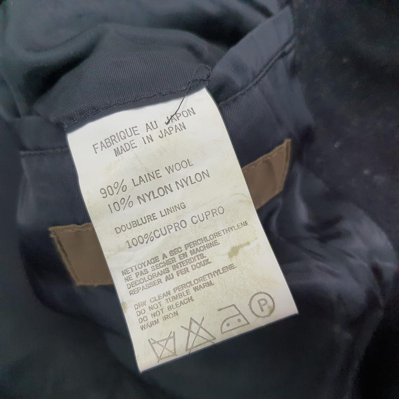 K&ouml;nnte beinhalten: Nahaufnahme eines Kleidungsst&uuml;cketiketts mit Text in Englisch und Franz&ouml;sisch. Das Etikett gibt die Stoffzusammensetzung an: 90 % Wolle, 10 % Nylon und 100 % Cupro-Futter. Es enth&auml;lt auch Pflegehinweise und den Text "Made in Japan."