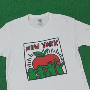 Puede incluir: Camiseta blanca con un dise&ntilde;o gr&aacute;fico que presenta una manzana roja con una hoja verde y las palabras "NEW YORK" en un recuadro con contorno negro. El dise&ntilde;o incluye figuras verdes y l&iacute;neas blancas radiantes.