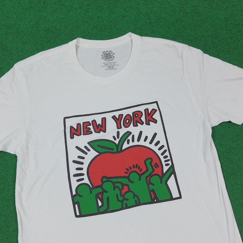 Puede incluir: Camiseta blanca con un dise&ntilde;o gr&aacute;fico que presenta una manzana roja con una hoja verde y las palabras "NEW YORK" en un recuadro con contorno negro. El dise&ntilde;o incluye figuras verdes y l&iacute;neas blancas radiantes.