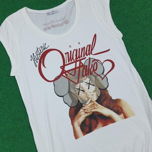 Puede incluir: Una camiseta blanca sin mangas con un estampado gr&aacute;fico de una persona con cabello largo casta&ntilde;o y una m&aacute;scara gris en forma de nube sobre los ojos marcada con X negras. El texto "Hysteric" y "Original Fake" aparece en rojo sobre la figura.