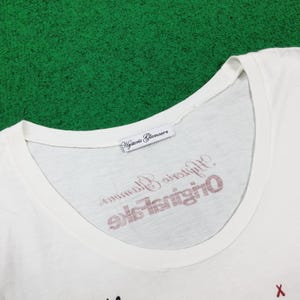 Puede incluir: Una camiseta blanca con cuello redondo ancho que presenta una etiqueta de tela blanca en el cuello interior con el texto Hysteric Glamour. El &aacute;rea del pecho muestra texto en rojo y negro que dice Hysteric Glamour Original Fake.