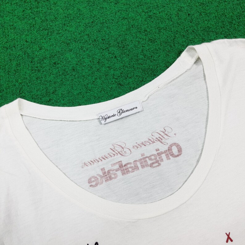Puede incluir: Una camiseta blanca con cuello redondo ancho que presenta una etiqueta de tela blanca en el cuello interior con el texto Hysteric Glamour. El &aacute;rea del pecho muestra texto en rojo y negro que dice Hysteric Glamour Original Fake.