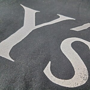 K&ouml;nnte beinhalten: Nahaufnahme eines dunkelgrauen Textils mit den wei&szlig;en Buchstaben "YS" in einer stilisierten Schriftart. Die Buchstaben scheinen auf den Stoff geschablont oder gedruckt zu sein, mit einem leicht verwitterten Aussehen. Die Textur des Stoffes ist sichtbar.