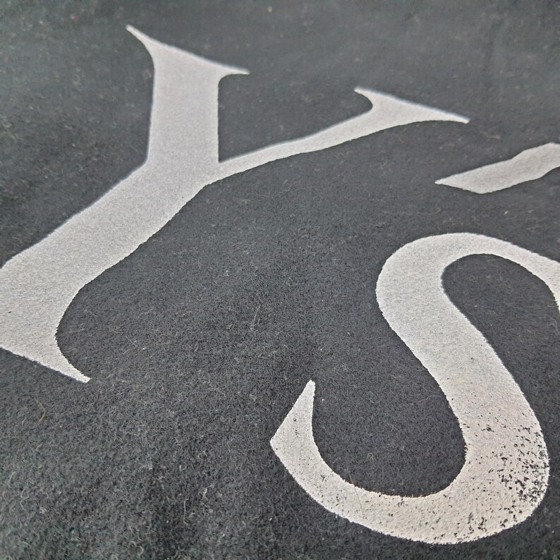 K&ouml;nnte beinhalten: Nahaufnahme eines dunkelgrauen Textils mit den wei&szlig;en Buchstaben "YS" in einer stilisierten Schriftart. Die Buchstaben scheinen auf den Stoff geschablont oder gedruckt zu sein, mit einem leicht verwitterten Aussehen. Die Textur des Stoffes ist sichtbar.