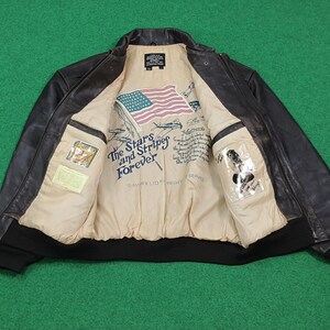 Puede incluir: Una chaqueta bomber de cuero marr&oacute;n oscuro con forro interior beige. El forro presenta una bandera estadounidense impresa, ilustraciones de aviones y el texto The Stars and Stripes Forever. Incluye un dobladillo y pu&ntilde;os de canal&eacute; negros.