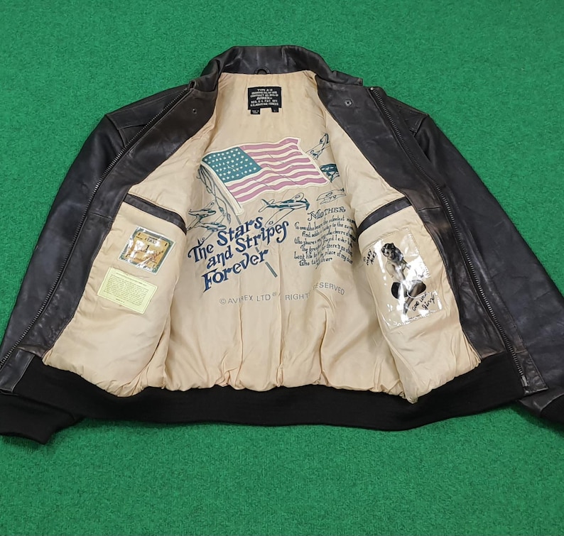 Puede incluir: Una chaqueta bomber de cuero marr&oacute;n oscuro con forro interior beige. El forro presenta una bandera estadounidense impresa, ilustraciones de aviones y el texto The Stars and Stripes Forever. Incluye un dobladillo y pu&ntilde;os de canal&eacute; negros.