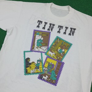 May include: White t-shirt featuring the text "TIN TIN" in black block letters above four colorful postage stamp-style illustrations. The illustrations depict scenes with characters and dogs.