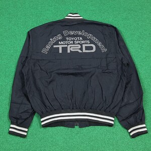 May include: Black nylon jacket with white and gray accents. The jacket features the text "Racing Development TOYOTA MOTOR SPORTS TRD" on the back. The collar, cuffs, and hem have white stripes.