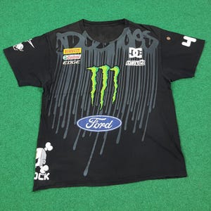 May include: A black short-sleeve t-shirt featuring a large green Monster Energy logo, a blue Ford oval emblem, and various sponsor logos including Pirelli, Castrol Edge, and DC Dirt 2. The design includes grey paint drip graphics and a cartoon character.
