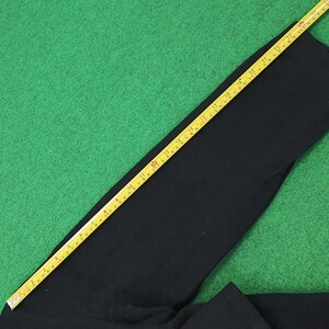 K&ouml;nnte beinhalten: Schwarzes Kleidungsst&uuml;ck, m&ouml;glicherweise eine Hose, auf einer gr&uuml;nen Oberfl&auml;che. Ein gelbes Ma&szlig;band liegt diagonal &uuml;ber dem Kleidungsst&uuml;ck und zeigt Messungen in Zentimetern an. Der Stoff hat eine dunkle, einfarbige Farbe.