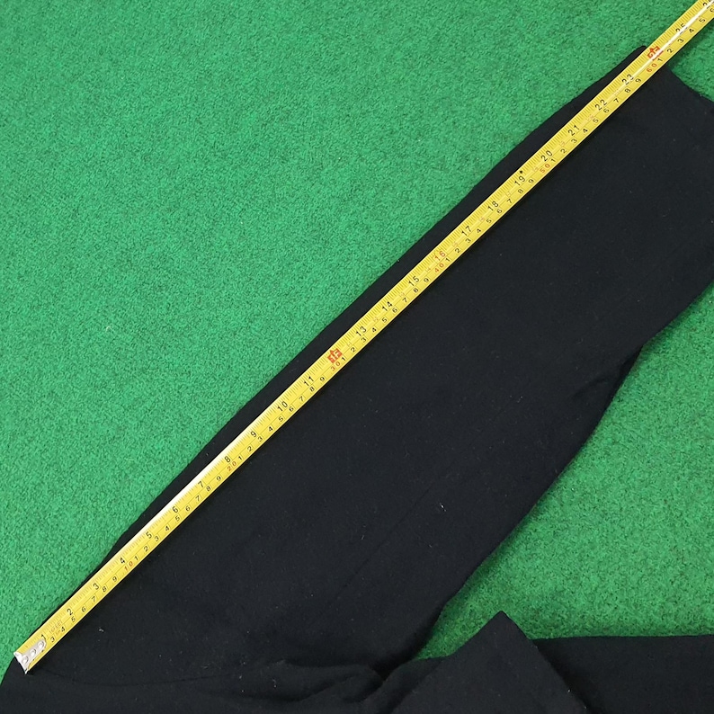 K&ouml;nnte beinhalten: Schwarzes Kleidungsst&uuml;ck, m&ouml;glicherweise eine Hose, auf einer gr&uuml;nen Oberfl&auml;che. Ein gelbes Ma&szlig;band liegt diagonal &uuml;ber dem Kleidungsst&uuml;ck und zeigt Messungen in Zentimetern an. Der Stoff hat eine dunkle, einfarbige Farbe.