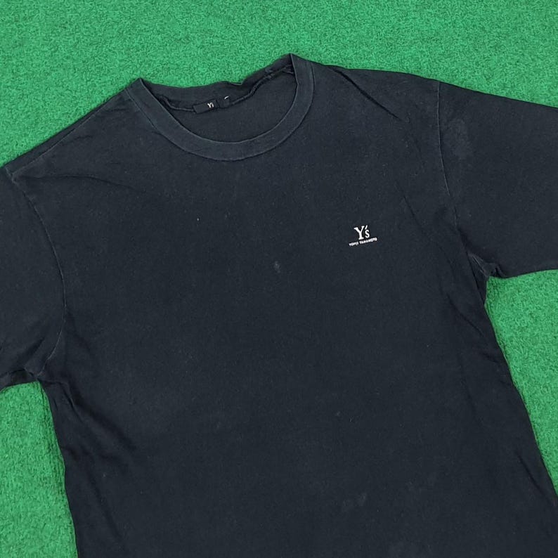 May include: A black short-sleeved t-shirt with a crew neckline. The shirt features a small white embroidered logo on the left chest that reads Y's Yohji Yamamoto. The fabric appears to have a slightly faded texture throughout the garment surface.
