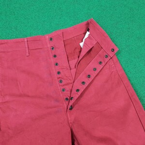 May include: Close-up of red pants with a button-up fly. The pants have a high waist and a straight leg design. The buttons are black and the fabric appears to be a sturdy cotton or blend. The pants are laid flat on a green surface.