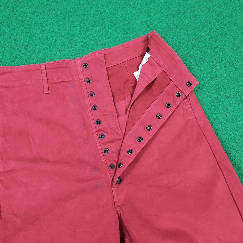 May include: Close-up of red pants with a button-up fly. The pants have a high waist and a straight leg design. The buttons are black and the fabric appears to be a sturdy cotton or blend. The pants are laid flat on a green surface.