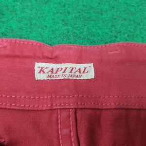 May include: Close-up of red fabric with a white label that reads "KAPITAL" and "MADE IN JAPAN". The fabric has visible stitching and a textured appearance. The background is a green surface.