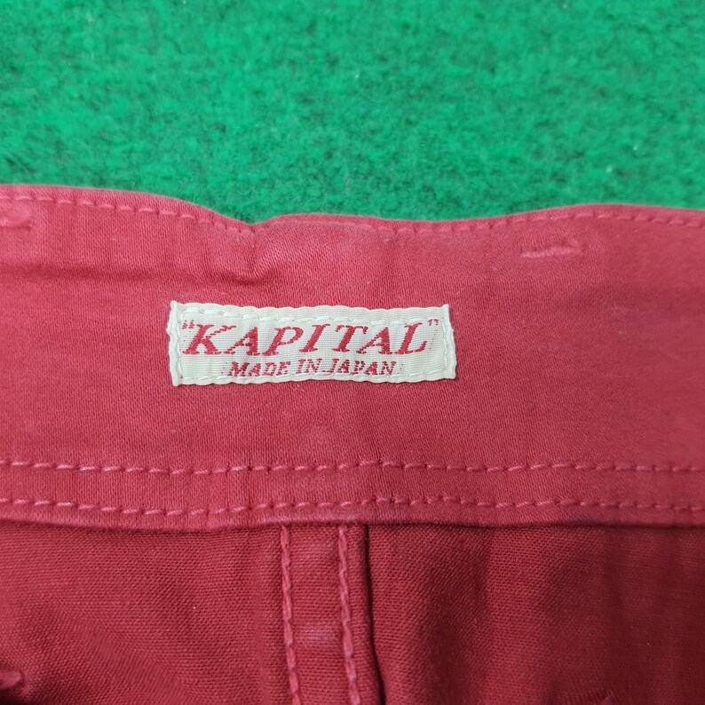 May include: Close-up of red fabric with a white label that reads "KAPITAL" and "MADE IN JAPAN". The fabric has visible stitching and a textured appearance. The background is a green surface.