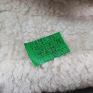 Puede incluir: Primer plano de una tela texturizada de color blanquecino. Una peque&ntilde;a etiqueta rectangular verde con texto negro es parcialmente visible, mostrando n&uacute;meros y letras. La tela parece suave y esponjosa.