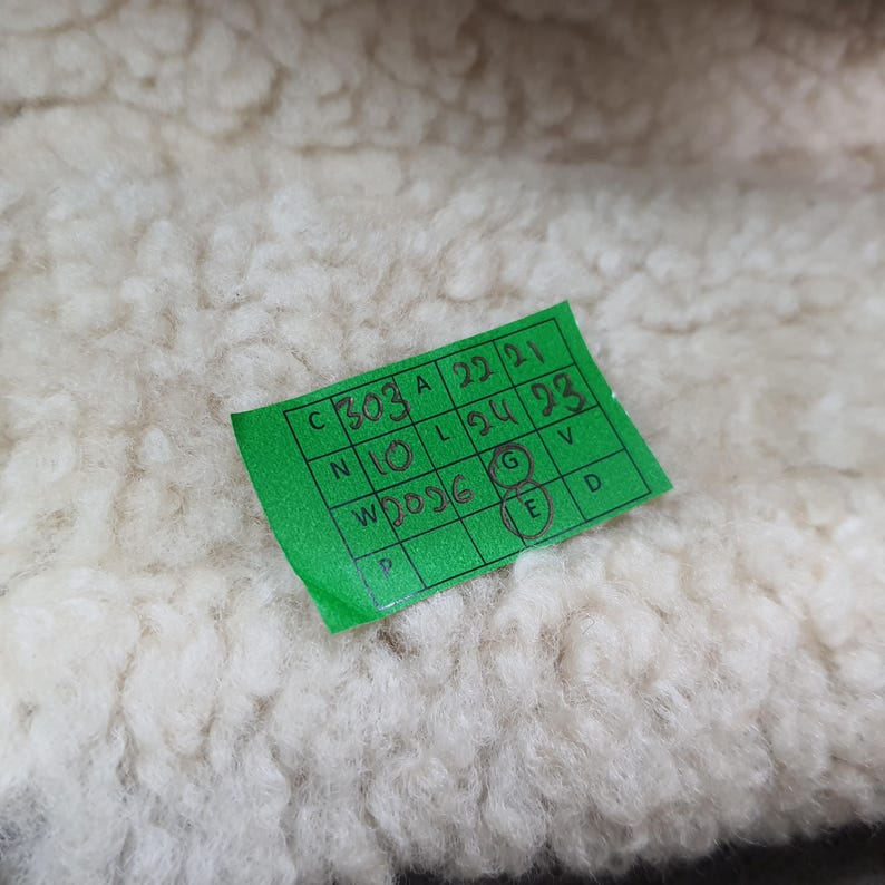 Puede incluir: Primer plano de una tela texturizada de color blanquecino. Una peque&ntilde;a etiqueta rectangular verde con texto negro es parcialmente visible, mostrando n&uacute;meros y letras. La tela parece suave y esponjosa.
