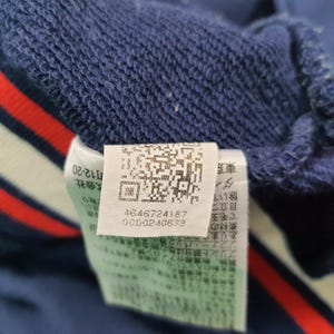 Puede incluir: Primer plano de una tela azul marino con un detalle de rayas blancas, rojas y azules. Una etiqueta blanca con un c&oacute;digo QR y texto est&aacute; adherida a la tela. La etiqueta incluye los n&uacute;meros 4646724187 y 0000240673.