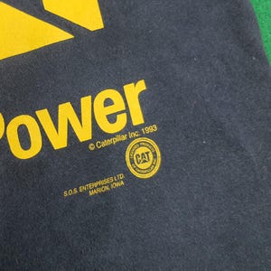 May include: A dark gray t-shirt featuring yellow screen-printed text that reads Power, Caterpillar Inc. 1993, and S.O.S. ENTERPRISES LTD. MARION, IOWA. A circular yellow and black CAT Licensed Product logo is printed below the word Power.