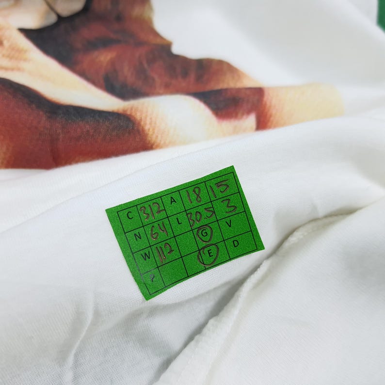 Puede incluir: Una camiseta blanca con un estampado gr&aacute;fico en marr&oacute;n y beige. Una etiqueta rectangular verde est&aacute; adherida a la tela, mostrando medidas escritas a mano que incluyen C 312, A 18, 15, N 64, L 30.5, 3, W 112, G y E, detallando las medidas de la prenda.