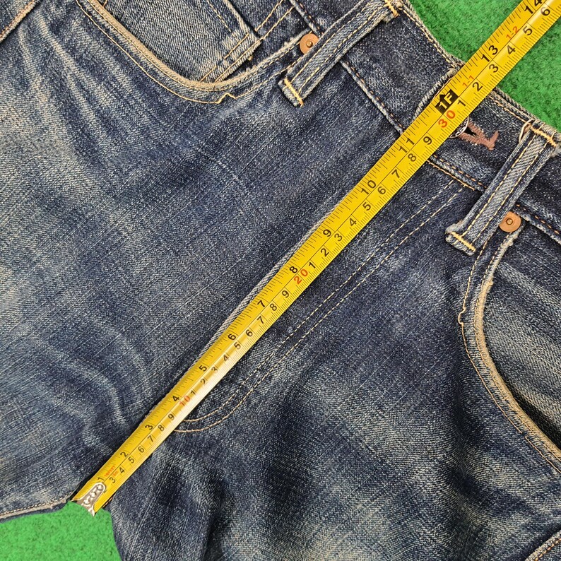 K&ouml;nnte beinhalten: Nahaufnahme einer blauen Jeansshorts mit einem gelben Ma&szlig;band &uuml;ber dem Stoff. Das Band zeigt Messungen in Zentimetern, die die Abmessungen des Kleidungsst&uuml;cks hervorheben. Die Shorts haben sichtbare N&auml;hte und klassische Taschendetails.