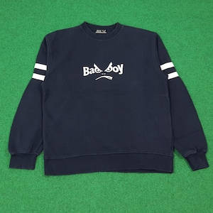 May include: Navy blue sweatshirt with the text "Bad Boy" in white across the chest. The sweatshirt has white stripes on the sleeves and a crew neck. The garment is displayed on a green surface.