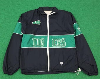 May include: A navy blue and green athletic jacket with the word "TIGERS" across the chest. The jacket has a white zipper and a small logo on the upper left chest. The sleeves have green accents and the jacket is on a green surface.
