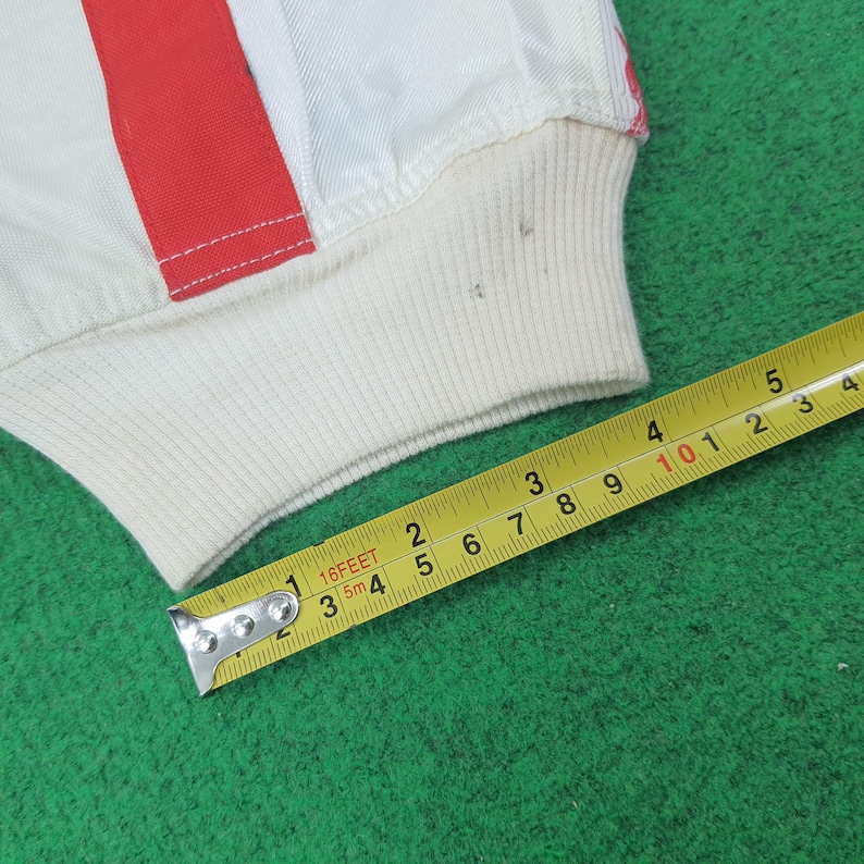 May include: Close-up of a white garment with a red stripe detail. The garment has a ribbed, off-white cuff. A yellow measuring tape shows measurements in inches and feet.