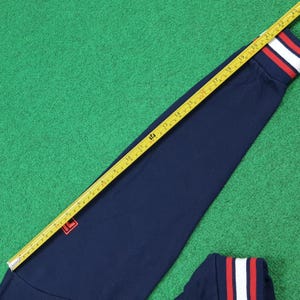 Puede incluir: Top deportivo azul marino de manga larga con pu&ntilde;os a rayas rojas y blancas. El top presenta un peque&ntilde;o logotipo rojo en el pecho. Una cinta m&eacute;trica amarilla se coloca en diagonal, mostrando medidas en cent&iacute;metros.