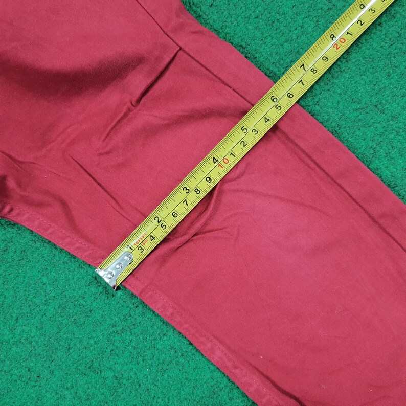 May include: Close-up of a pair of red pants with a yellow measuring tape. The tape shows measurements in inches. The pants are on a green surface.