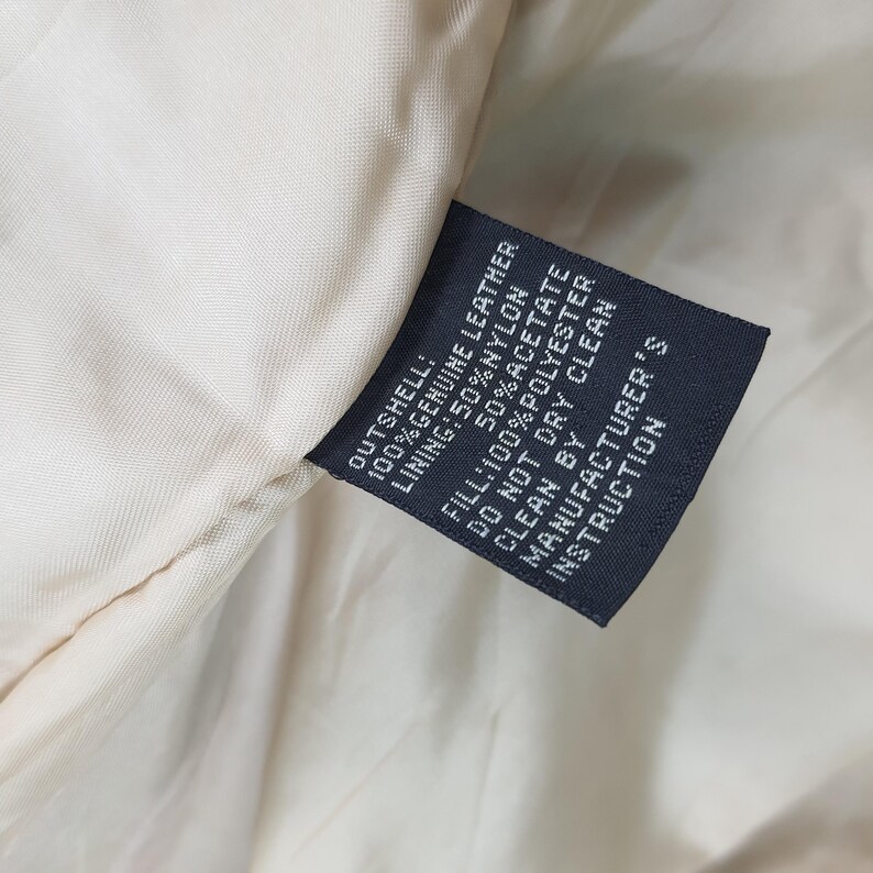 Puede incluir: Una etiqueta de cuidado rectangular de color azul oscuro cosida en un forro de tela color crema. El texto dice: OUTSHELL: 100% GENUINE LEATHER, LINING: 50% NYLON 50% ACETATE, FILL: 100% POLYESTER, DO NOT DRY CLEAN, CLEAN BY MANUFACTURER'S INSTRUCTION.