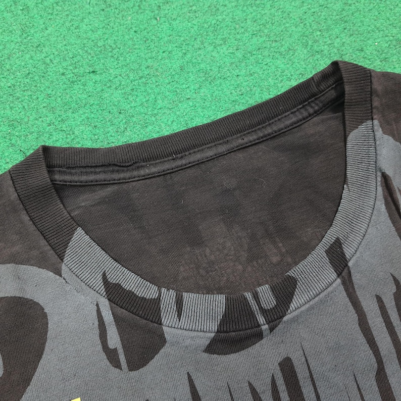 May include: A black crew neck t-shirt featuring a dark gray abstract graphic pattern across the fabric. The garment has a ribbed collar and short sleeves, with the design extending from the neckline down across the front of the shirt.