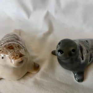 May include: Two ceramic seal figurines. One is light beige with brown spots, the other is grey with darker grey spots. Both seals are lying down with their flippers extended.