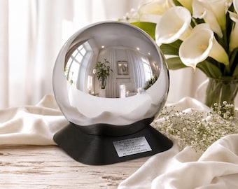 May include: A reflective silver sphere memorial on a black base. The sphere reflects a room with white curtains, a plant, and furniture. A small plaque on the base reads "FOREVER IN OUR HEARTS". White calla lilies and baby's breath are in the background.