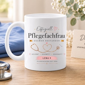 Könnte beinhalten: Eine weiße Keramik-Tasse mit schwarzem und rosa Text, der Offiziell. Pflegefachfrau EXAMEN BESTANDEN lautet, ergänzt durch eine goldene Stethoskop-Grafik, den Namen LENA sowie die Aufschriften GELERNT GEKÄMPFT GESCHAFFT und PFLEGEFACHFRAU SEIT 2026.