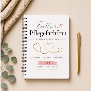 Könnte beinhalten: Ein weißes Notizbuch mit Spiralbindung und dem Text Endlich. Pflegefachfrau, Examen bestanden, Gelernt, Gekämpft, Geschafft, Lena sowie Pflegefachfrau seit 2026. Das Cover zeigt ein goldenes Stethoskop in Herzform und kleine rosa Herzen.