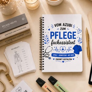 Könnte beinhalten: Ein weißes Spiralnotizbuch mit blauem und schwarzem Text, der VOM AZUBI ZUM PFLEGE fachassistent, STOLZ KOMPETENT MIT HERZ und ZUKUNFT GESTALTEN lautet, ergänzt durch Illustrationen eines Stethoskops, eines Herzens und eines blauen Kasacks.