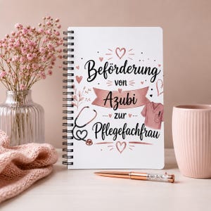 Könnte beinhalten: Ein weißes Spiralnotizbuch mit dem schwarzen Text "Beförderung von Azubi zur Pflegefachfrau", roségoldenen Akzenten, einer Stethoskop-Illustration und einer Grafik eines Kasacks, verziert mit kleinen Herzen und floralen Elementen.