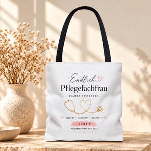 Könnte beinhalten: Eine weiße Tragetasche mit schwarzen Henkeln, bedruckt mit dem Text Endlich. Pflegefachfrau, Examen bestanden, Gelernt, Gekämpft, Geschafft, dem Namen Lena auf einem rosa Pinselstrich und dem Text Pflegefachfrau seit 2026 mit einem goldenen Stethoskop-Herz.