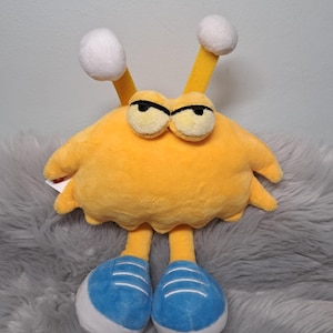 May include: A plush toy shaped like a yellow creature with a rounded body, two antennae topped with white spheres, and two large eyes with black pupils. The creature has two yellow legs ending in blue sneakers with white laces and white soles.
