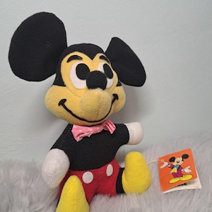 Pu&ograve; includere: Un peluche vintage di Topolino con orecchie nere, viso giallo e corpo nero. Il giocattolo indossa pantaloncini rossi, piedi gialli e un papillon rosa. &Egrave; attaccata una piccola etichetta con un'illustrazione di Topolino.