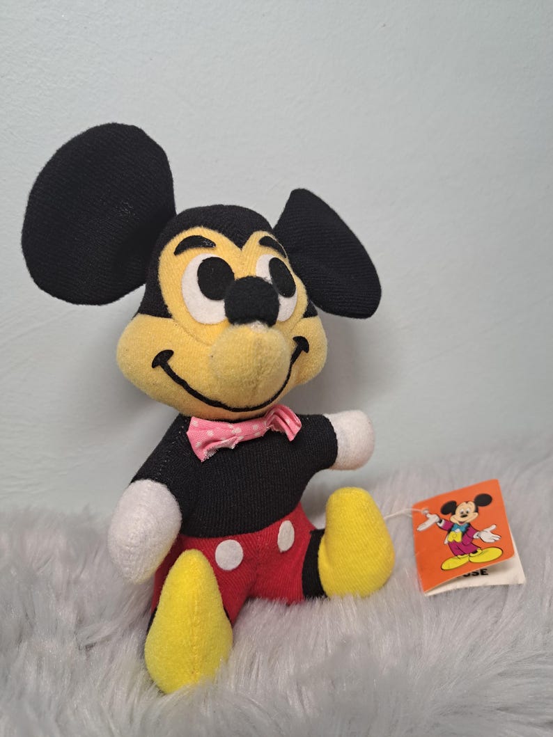 Pu&ograve; includere: Un peluche vintage di Topolino con orecchie nere, viso giallo e corpo nero. Il giocattolo indossa pantaloncini rossi, piedi gialli e un papillon rosa. &Egrave; attaccata una piccola etichetta con un'illustrazione di Topolino.