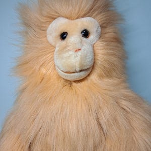 May include: A plush monkey toy with a light brown, shaggy fur. The monkey has a light tan face, with black eyes and a smiling mouth. The toy is designed to resemble a monkey.