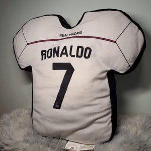 Puede incluir: Un coj&iacute;n con forma de camiseta blanca con bordes negros que presenta el texto REAL MADRID, el nombre RONALDO y el n&uacute;mero 7 en letras negras. El dise&ntilde;o incluye una fina l&iacute;nea horizontal roja en el pecho y un peque&ntilde;o logotipo en la base.